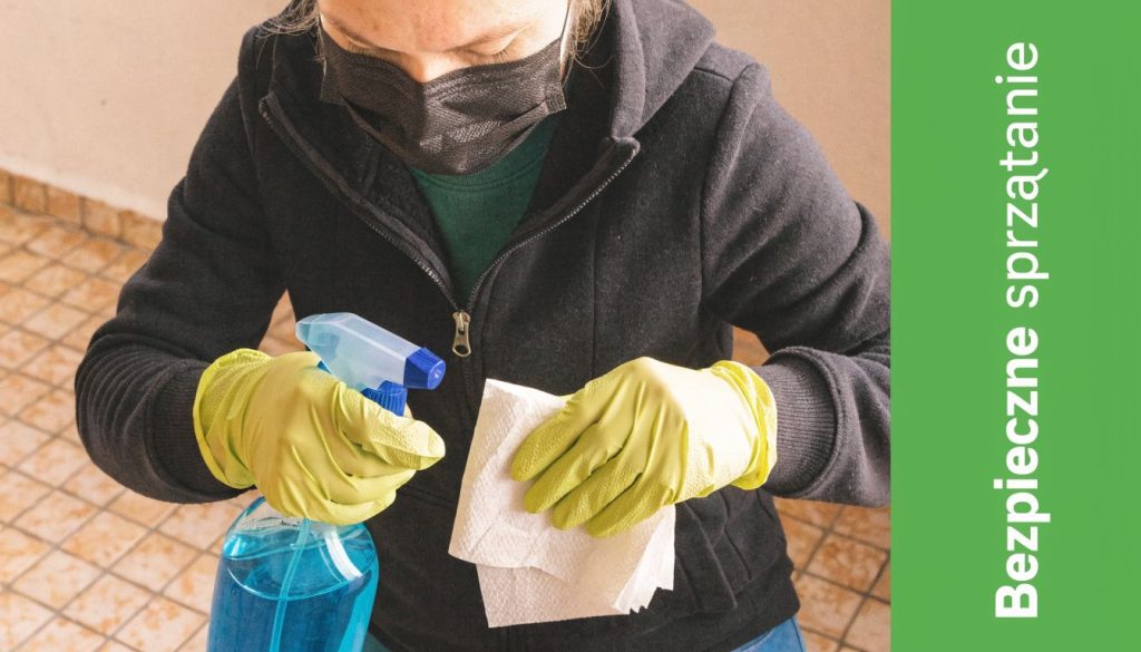 Osoba w masce ochronnej i żółtych rękawiczkach czyści powierzchnię płynem w sprayu oraz ręcznikiem papierowym.